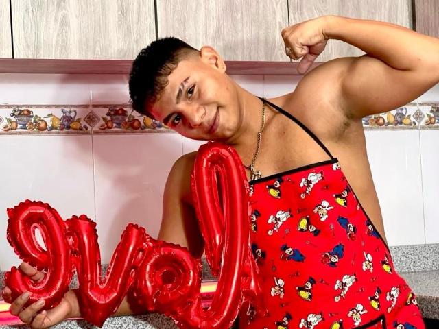 RichiMontes, com uma estrutura muscular, cabelo preto curto e pele bronzeada, vestindo um avental vermelho com estampas do Mickey Mouse, exibindo o braço enquanto segura balões vermelhos com a inscrição "Swoo"..