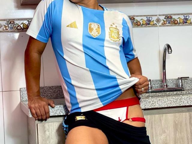 RichiMontes, gekleed in een blauw en wit Argentijns voetbalshirt, trekt zwarte shorts omhoog om rode bandjes van wit ondergoed te laten zien, waarbij een rode string zichtbaar wordt, met een wastafel op de achtergrond..