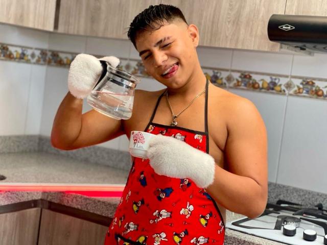 RichiMontes, con una complexión robusta, morena y musculosa, lleva un delantal rojo con estampados de dibujos animados, guantes blancos y un collar, y sonríe mientras vierte agua de un vaso..