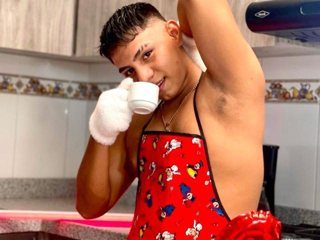 RichiMontes com o cabelo molhado, avental vermelho com estampas de desenhos animados, segurando uma xícara branca, sorrindo, levantando o braço, mostrando a axila, cozinha, usando uma luva branca felpuda..
