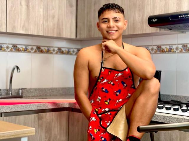 RichiMontes, muscoloso, con la pelle abbronzata, capelli castani corti, indossa un grembiule rosso con motivi di farfalle, è seduto sul bancone della cucina, con la gamba destra sollevata, sorridendo, dietro di lui c'è una stufa nera, la mano sinistra è appoggiata al mento..