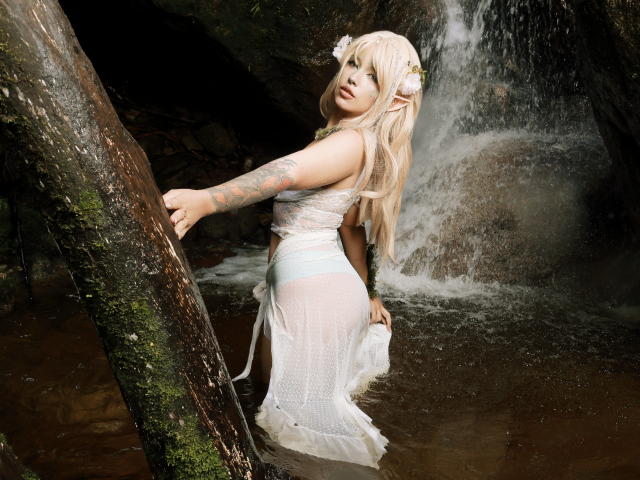 CarlaMiceli with long blonde hair, elf ears, white lace dress, and floral headpiece, standing in a waterfall, showing her tattooed arm and pale blue panties, holding a tree.