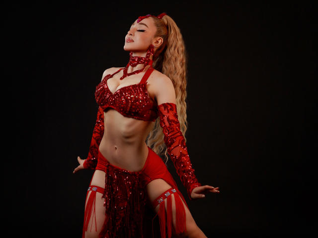 AnastasiaKovak, with long blonde ponytail, wearing red sequined bra and skirt, red arm sleeves, and thigh straps, stands against dark background, eyes closed, hands on hips, smiling.