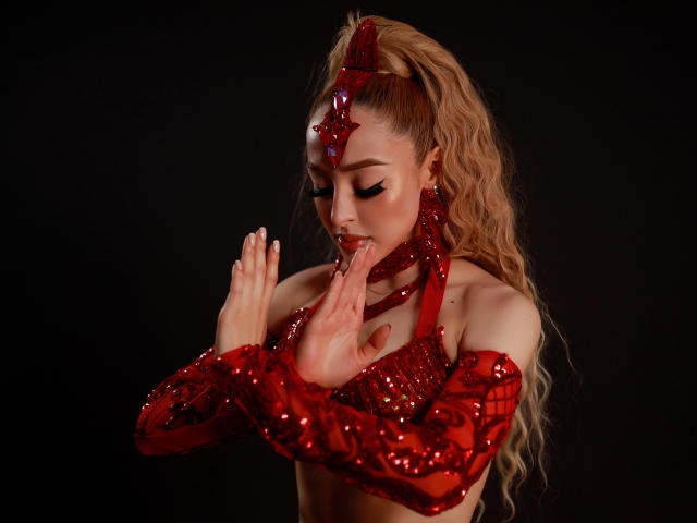 AnastasiaKovak with red sequined arm covers, long wavy blonde hair, and a red headpiece, licking her finger, eyes closed, hands near her face, black background.