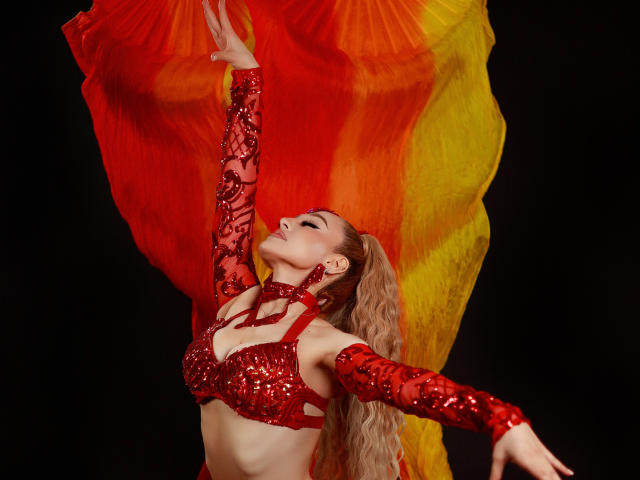 AnastasiaKovak with red sequined bra and arm sleeves, blonde ponytail, eyes closed, arms raised, holding red-yellow-orange fan, black background, posing seductively, showing her tits.