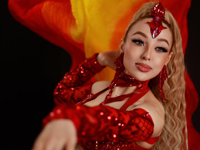 AnastasiaKovak with long blonde hair, red sequin dress, and matching red headpiece, exposing her shiny red-tipped nipples, has a sexy, confident look, and is pointing forward.