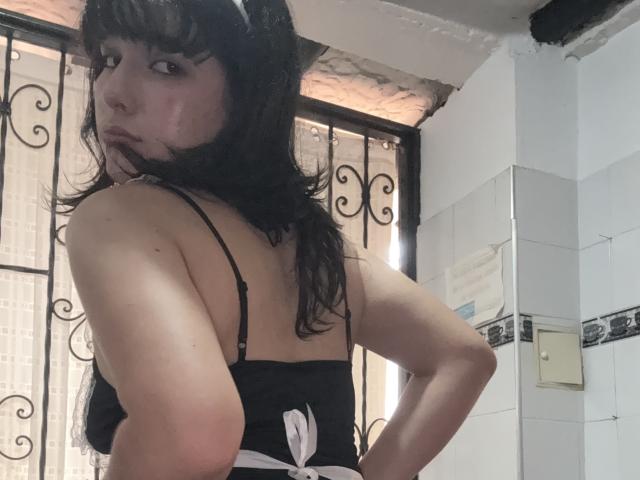HairyValeri with wet black hair, looking back, wearing a black spaghetti-strap top, showing her bare back and white apron, in a white-tiled room with a metal door.