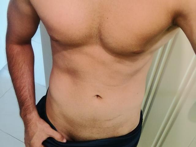 EliasX's bare, muscular torso with visible abs, light skin, and a slight tan. EliasX's right hand rests on black pants, exposing a small navel and faint pubic hair.