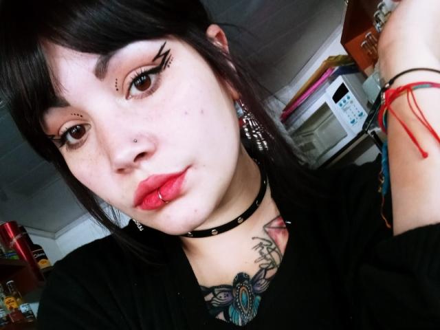SweetAmeliee mit schwarzen Haaren, Eyeliner, Nasenring, rotem Lippenring, schwarzem Choker, Tattoos und einem roten Armband, in einem Selfie, das ihre Piercings und ihren Gothic-Stil zeigt..