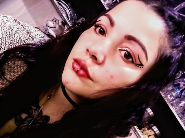 SweetAmeliee, con i capelli scuri, le labbra rosse carnose, una collana nera e l'eyeliner con tre piccoli punti neri sopra l'occhio destro, guarda l'obiettivo. Il suo viso è leggermente inclinato..