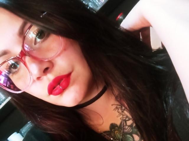 SweetAmeliee mit roten Lippen, Brille, schwarzem Choker und Tattoo, die ihre Brüste in einem Selfie zur Schau stellt, dunkles Haar und ein süßes, sexy Gesicht..