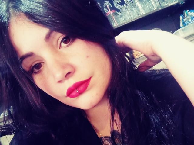 SweetAmeliee mit dunklem Haar, rotem Lippenstift und einem leichten Lächeln, die ihre Haare berührt. Sie hat einen kleinen Muttermal auf ihrer linken Wange und trägt ein schwarzes Oberteil..