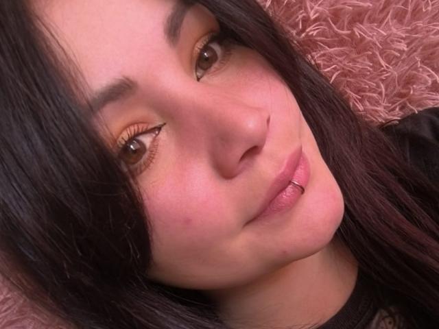 Le visage de SweetAmeliee, avec ses yeux marron et ses longs cils, ses cheveux noirs et lisses, sa peau rosée et son piercing au menton, est allongé sur une surface rose et moelleuse..
