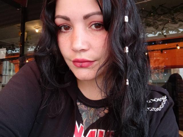 SweetAmeliee heeft donker, golvend haar, rode lippenstift en een kleine piercing in haar lip. Ze draagt een zwart shirt waarbij een tatoeage op haar nek zichtbaar is. Haar ogen zijn bruin..
