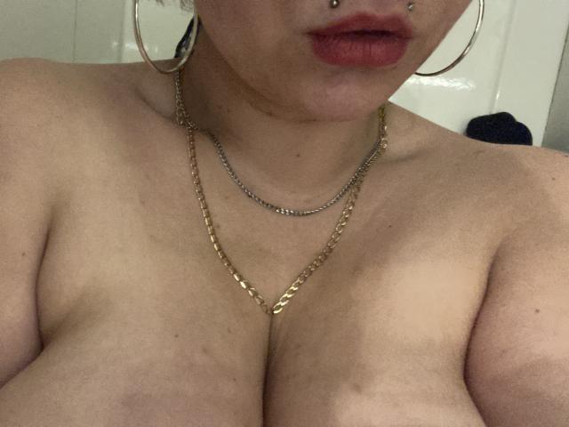 Les seins volumineux de PixiiieKitty sont visibles, elle porte un collier en or et des boucles d'oreilles cerceaux, avec du rouge à lèvres et un piercing au nez, sur une photo prise dans une faible luminosité, légèrement floue..