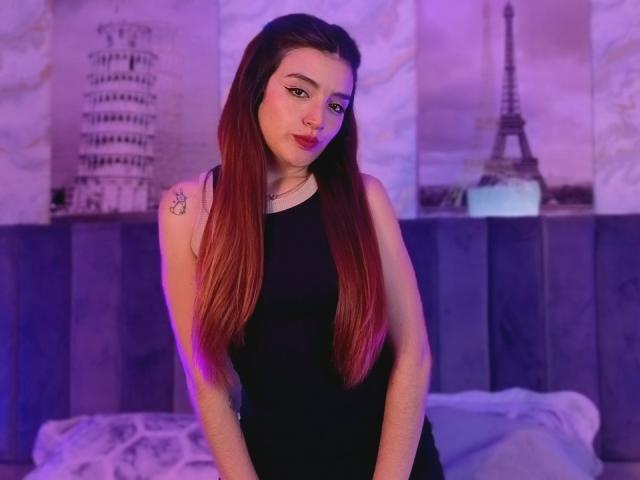 ArianaWithe, with long red-brown hair, black sleeveless top, and sun tattoo on right shoulder, stands in purple-lit room, looking at camera, hands on her thighs, slight smile.