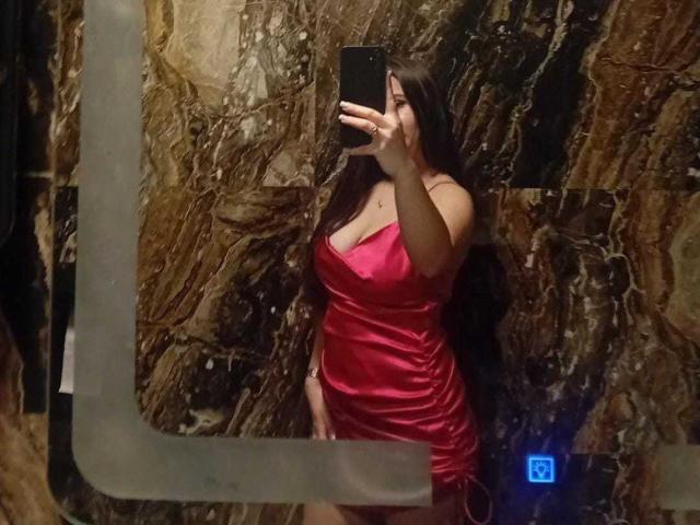 DarkSweettie se prend en photo devant un miroir, vêtue d'une robe rouge en satin, mettant en valeur ses seins volumineux et ses courbes, avec de longs cheveux bruns, tout en tenant un téléphone noir..
