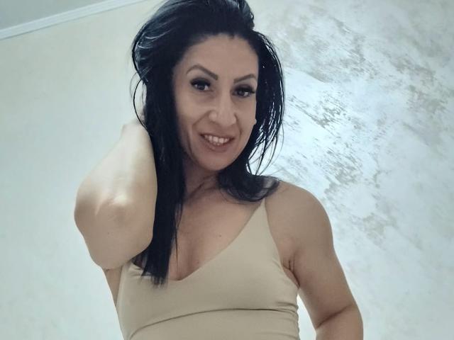 LanceDance, sonriendo, con el cabello negro y mojado, vistiendo una camiseta sin mangas beige, mostrando sus pechos, con el brazo levantado, mirando hacia abajo a la cámara, en una habitación luminosa..