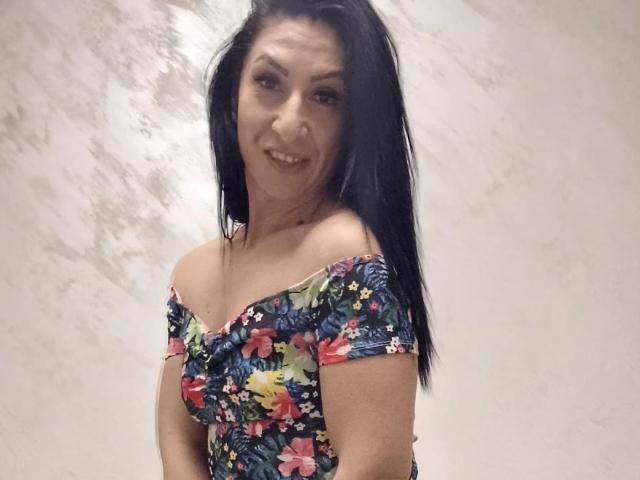 LanceDance met zwart haar, een bloemenjurk met open schouder, middelgrote borsten, lachend, kijkt over haar schouder, lichte huid, lichtelijk rommelig haar, staat tegen een effen muur, nonchalante pose, medium shot..