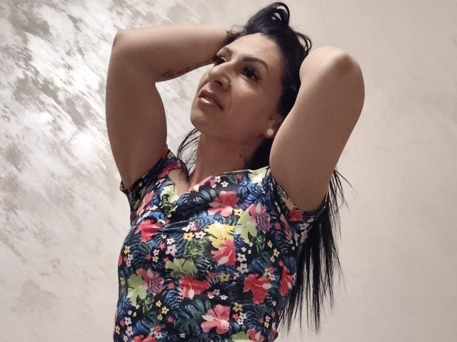 LanceDance con le braccia alzate, capelli neri, sorridente, indossa una camicia colorata con fiori, mostra piccoli seni, fianchi di media grandezza, tatuaggio sul braccio sinistro, in piedi di fronte a una parete bianca con una texture..