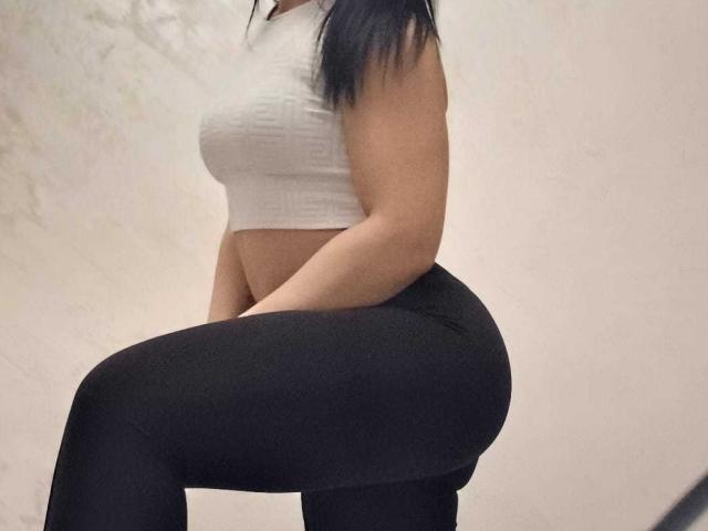 LanceDance, con i capelli neri, indossa un top corto bianco e pantaloni neri aderenti, mostrando i suoi fianchi formosi e i piccoli seni, con il ginocchio piegato, vista laterale, in una stanza semplice..
