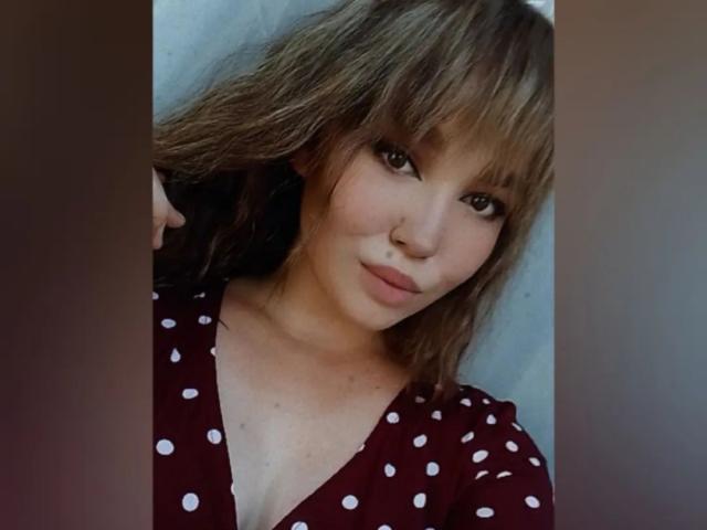 CoriNita with wavy brown hair, bangs, and brown eyes, wearing a red polka dot top, showing a subtle smile, and holding her hair on the left side.