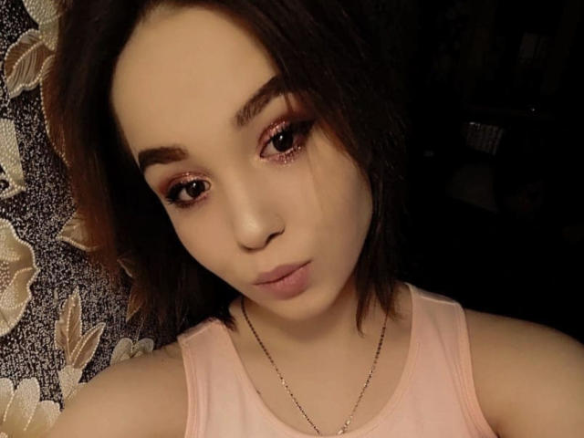 CoriNita with light skin, brown eyes, and pink makeup, wearing a pink tank top, has short brown hair, and a necklace, looking at the camera with a neutral expression.