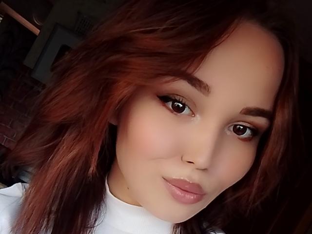 CoriNita with red, wavy hair, brown eyes, and glossy pink lips. Her white top contrasts with her fair skin. She's looking at the camera with a subtle smile.