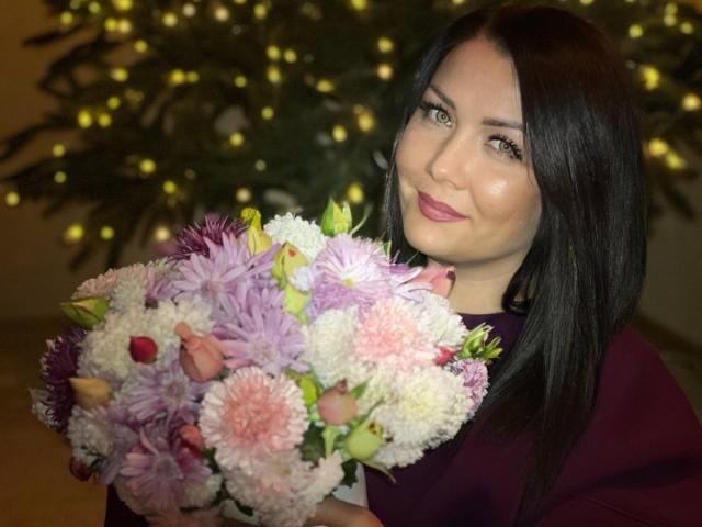 LanaMoon69 with long black hair, wearing a purple shirt, holding a bouquet of pink, white, and purple chrysanthemums, smiling at the camera. Christmas tree with lights in background.