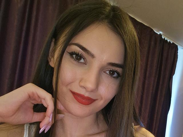 MiaAddison with long brown hair, red lipstick, and eyeliner, smiling at the camera, hand near face, purple curtain in background, light skin, brown eyes, pink nails.