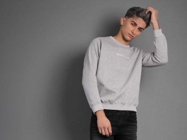 CandyEngel, standing, gray sweatshirt, black jeans, right hand in hair, short gray hair, serious expression, gray background, slim build, slightly leaning to left.