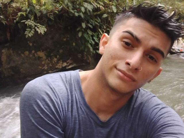 CandyEngel with short, dark hair, a nose ring, and a subtle smile, wearing a gray shirt, standing by a flowing river, looking slightly to the side.