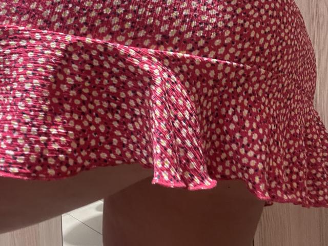 O vestido floral vermelho e branco de Barbarica, com saia evasê, ligeiramente levantado, revelando seu bumbum nu e bronzeado. O tecido tem textura, com um padrão de pequenas flores..