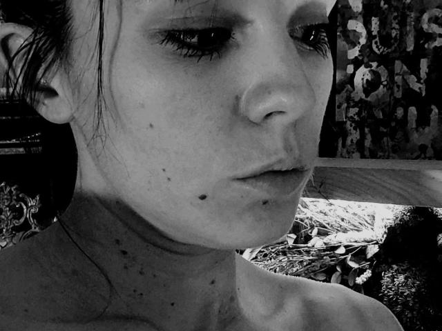 black-and-white photo of OrianaLaFrancaise with freckles and a small mole on her chin, half-closed eyes, and dark, slightly messy hair, looking downward with a neutral expression.