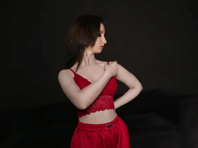 FlawLessLife with pale skin and brown hair, wearing a red lace bra and matching red pants, holds her bra strap, showing small tits and a slim, sexy figure.