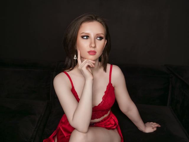 FlawLessLife with pale skin, brown hair, red lace bra, and matching skirt, sitting on black couch, hand near mouth, looking to the side, wearing triangle earrings, red lipstick.