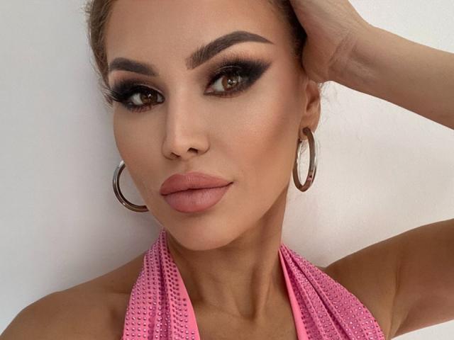 RadmilaQueen has dark, thick eyebrows, brown eyes with black eyeliner, pink lipstick, and wears large silver hoop earrings with a pink, textured halter top.