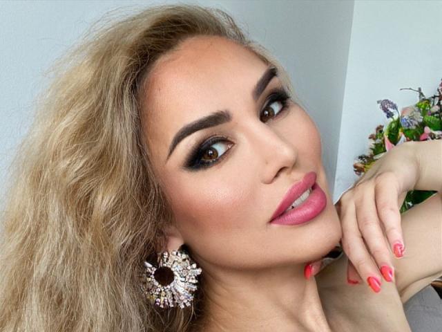 RadmilaQueen has blonde hair, brown eyes, pink lipstick, and wears diamond earrings. Her pink nails and subtle makeup highlight her sexy smile. She's leaning on a white wall.