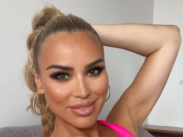 RadmilaQueen with tan skin, blonde ponytail, hoop earrings, brown eyes, thick eyebrows, long lashes, pink lipstick, and a pink top, looking at the camera with a slight smile.