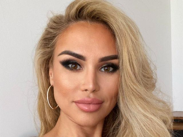 RadmilaQueen has blonde hair, brown eyes with thick black eyeliner, and pink lipstick. She's wearing gold hoop earrings and has a slightly tanned, smooth complexion.