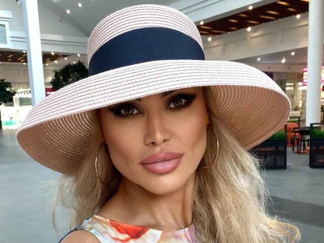 RadmilaQueen with a big, tan, straw hat, black band, and blonde hair, wearing a colorful dress, hoop earrings, and pink lipstick, looking straight at the camera.