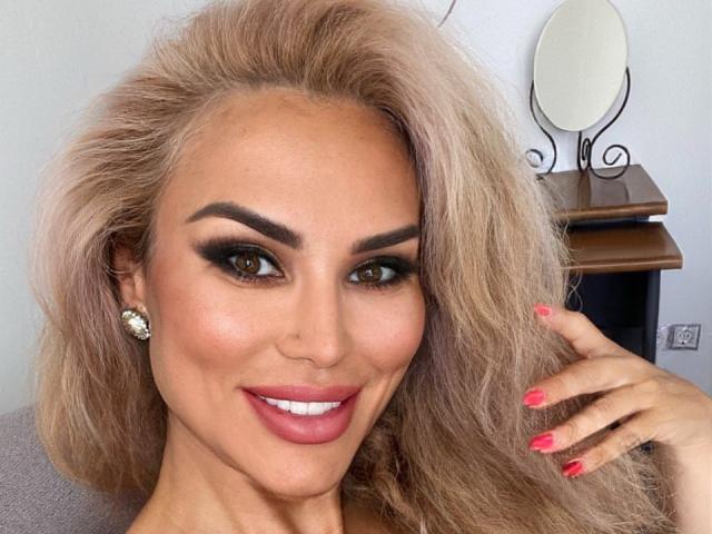 RadmilaQueen with blonde, wavy hair, brown eyes, heavy makeup, pink lipstick, pink nails, and diamond earrings, smiling at the camera. Her tits are slightly visible.