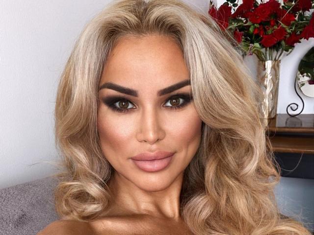 RadmilaQueen has blonde, wavy hair, brown eyes, thick eyebrows, and full pink lips. She's wearing dark, smoky eye makeup. Her tits are slightly visible, with a tan skin tone.