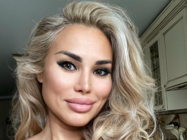 RadmilaQueen has blonde, wavy hair, full eyebrows, dark eyeliner, and pink lipstick. She's smiling with a slight pout, showing off her pretty brown eyes and smooth skin.