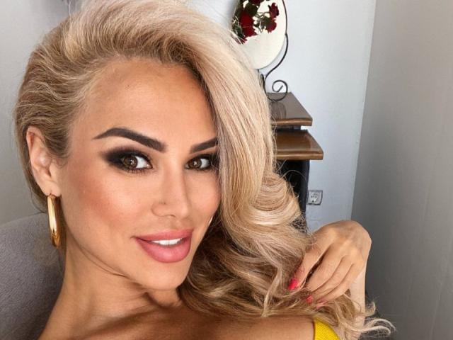 RadmilaQueen with blonde hair, heavy makeup, and pink lipstick, smiling, wearing gold earrings, and yellow top, showing red nails, and holding her wavy hair.