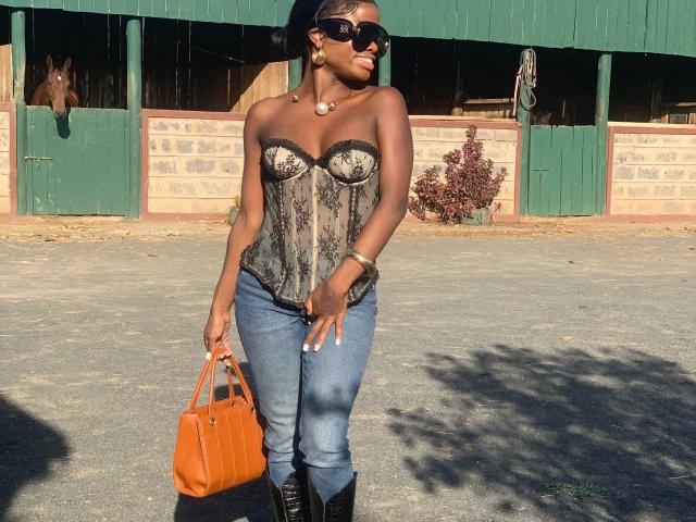 Essyreina69-hot with dark skin, wearing a black lace strapless corset, blue jeans, black boots, orange bag, and sunglasses, standing outdoors, holding a brown bag in her right hand.