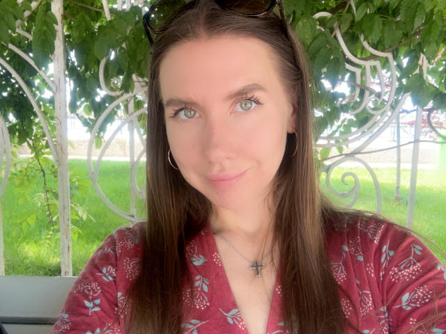 SannyManny has green eyes, long brown hair, and a slight smile. She's wearing a red floral shirt, a cross necklace, and black sunglasses on her head.