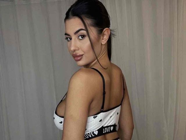 LatinaLoco with dark hair in a bun, wearing a white heart-patterned bra, showing off her tits, looking over her shoulder with a slight smile.