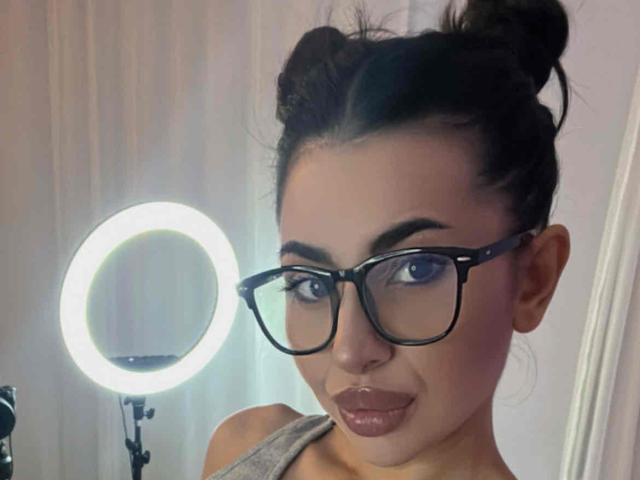 LatinaLoco with glasses, dark hair in a messy bun, smoky eye makeup, and glossy lips, smiling slightly, ring light on the left.