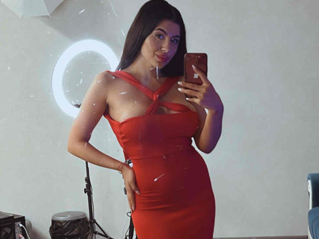 LatinaLoco with long dark hair, wearing a tight red dress with cutout chest, holding a phone, showing off her curvy ass and big boobs in a mirror selfie.